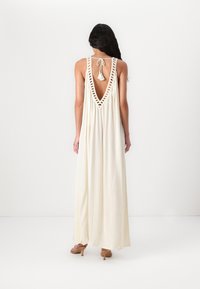 Robe longue crème avec un dos en V plongeant, detailing en dentelle le long de l'encolure et une fermeture à nouer. Tissu fluide tombant jusqu'aux chevilles.