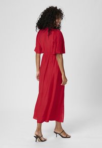 Robe maxi rouge fluide avec des manches courtes à volants, taille cintrée et texture douce. Le modèle porte des talons noirs à lanières.