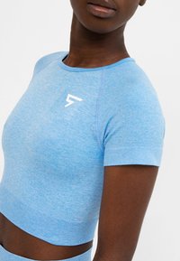 Detailní záběr osoby v světle modrém sportovním crop topu s krátkým rukávem a bílým logem na hrudi na bílém pozadí.