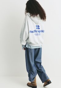Felpa grigia con cappuccio e logo blu, indossata sopra una maglietta a righe verdi e bianche. Pantaloni a righe blu larghi e stivaletti marroni alla caviglia.