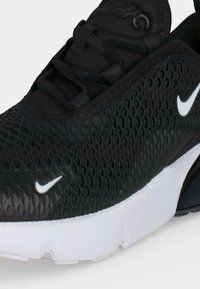 Nærbillede af sort Nike-sneaker med tekstureret overdel, sorte snørebånd, hvide mellemsåler og to hvide Nike swoosh-logoer synlige.