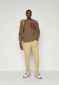 Καφέ ριχτό πουκάμισο με μακριά μανίκια και κόκκινη τσέπη στο στήθος, σε συνδυασμό με μπεζ παντελόνι και ανοιχτόχρωμα αθλητικά παπούτσια. Το μοντέλο κρατά μια κίτρινη τσάντα πλάτης.