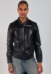 Veste bomber en cuir noir avec col et poignets côtelés, fermeture éclair et détail de marque à l'avant. Portée sur un pantalon en denim bleu clair.