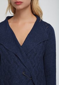 Navy blue knitted cardigan with a wide collar, textured wave pattern, and a decorative pin closure. Soft, medium-weight fabric.