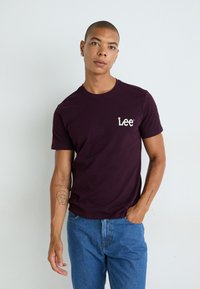 Bordeaux bomulds-t-shirt med et hvidt "Lee" logo på venstre bryst og en klassisk rund halsudskæring, parret med blå denim jeans.
