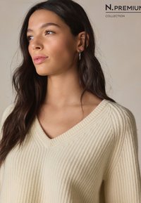 Krémsveter so sieťovaným vzorom a výstrihom do V, s vertikálnou rebrovanou textúrou a voľným strihom. Zvýraznený čistým lemom výstrihu.