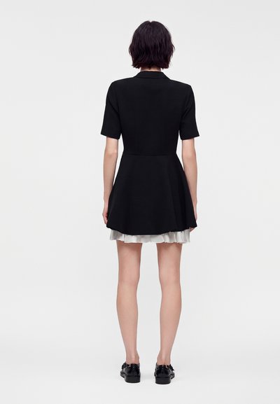 Femme debout de dos, portant une robe courte noire à manches courtes avec un ourlet de jupe blanc visible, associée à des chaussures noires sur fond blanc.