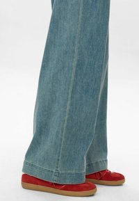 Világoskék, bő szárú farmernadrág sima textúrával, látható varrással és tiszta szegéllyel. Piros cipővel, fűzővel párosítva.