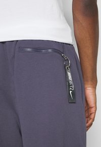 Nike-housut tummanharmaana, joissa on vetoketjullinen tasku metalliosilla ja tagi, jossa on Nike-logo ja brändäys.