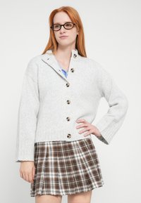 Jasnoszary dziany kardigan z wysokim kołnierzem, guzikami z przodu i szerokimi rękawami, zestawiony z brązową spódnicą w kratę. Tkanina o fakturze.