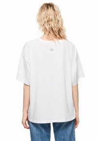 T-shirt bianca oversize con maniche corte, con scollatura rotonda e una piccola etichetta sulla parte superiore della schiena. Indossata con jeans azzurri chiari.