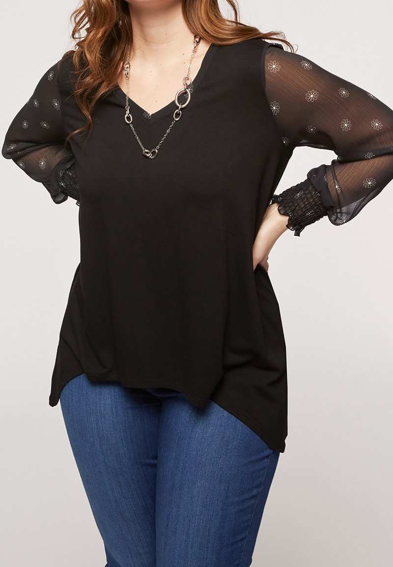 Black top with a V-neck and sheer, embroidered floral sleeves. Features a relaxed fit and textured cuffs, paired with blue jeans.