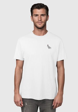 Mężczyzna ubrany w gładką białą koszulkę z małym nadrukiem gołębia w paskowanym szaliku na lewej stronie klatki piersiowej.