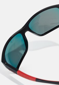 Solski briller med svart ramme og grønnfargede linser. Røde detaljer på stengene med en teksturert overflate. Lett og sporty design.
