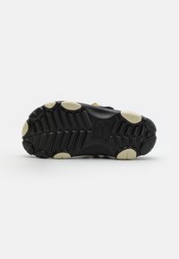 Suela de zapatilla negra con agarre de goma, patrón texturizado y acentos en crema. Presenta formas de amortiguamiento e indentaciones para mayor tracción.