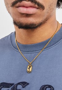 Gullkjede med en rektangulær anheng som har et silhuett-design, båret over en blå genser med rund hals.