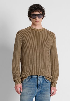 Giovane uomo che indossa un maglione marrone oversize in maglia, jeans blu e grandi occhiali da sole neri, in piedi davanti a uno sfondo chiaro e uniforme.