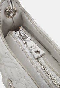 Intérieur d'un sac à main matelassé gris avec une fermeture éclair argentée ornée d'une découpe en forme de cœur et d'une étiquette de marque gravée. Texture en cuir lisse.