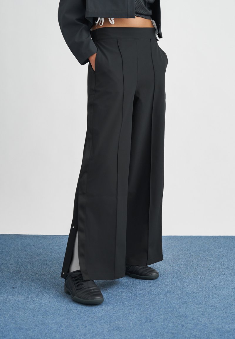 Μαύρο παντελόνι με φαρδιά μπατζάκια και λεία υφή, με πιέτες μπροστά και πλαϊνές σχισμές, συνδυασμένο με μαύρα αθλητικά παπούτσια.