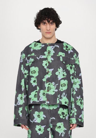 Veste en denim florale avec une base noire et de grandes fleurs vertes, deux poches poitrine et des boutons contrastés. La veste a une coupe carrée.