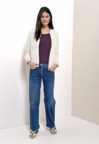 Blouson aviateur gris clair texturé porté sur un haut violet foncé, associé à un jean bleu coupe droite et des baskets avec des accents verts.