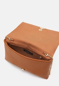 Bolso de piel de color marrón claro con cierre a presión en tono dorado, correa de cadena y textura suave. El interior cuenta con un forro de tela y un bolsillo con cremallera.