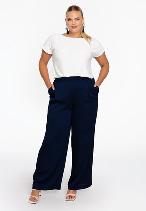 Weißes Kurzarmoberteil kombiniert mit navyweiten Beinhosen mit hohem Bund und Seitentaschen, gestylt mit weißen Riemchen-Heels.