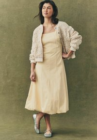 Svetlo rumena A-linijska obleka, teksturiran kremni kardigan s krošnjo in srebrni baletni čevlji s podrobnostmi trakova. Preprost in eleganten dizajn.