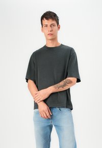 Czarnoszary, oversize t-shirt z okrągłym dekoltem, krótkimi rękawami i surowym wykończeniem, zestawione z jasnoniebieskimi dżinsami. Na ramieniu widoczny floralny tatuaż.