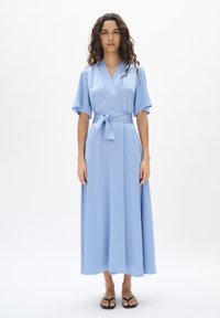 Helles blaues Wickelkleid mit kurzen, schwingenden Ärmeln, einem gebundenen Gürtel und einem bodenlangen Rock aus weichem, strukturiertem Stoff. Sandalen sind unten sichtbar.