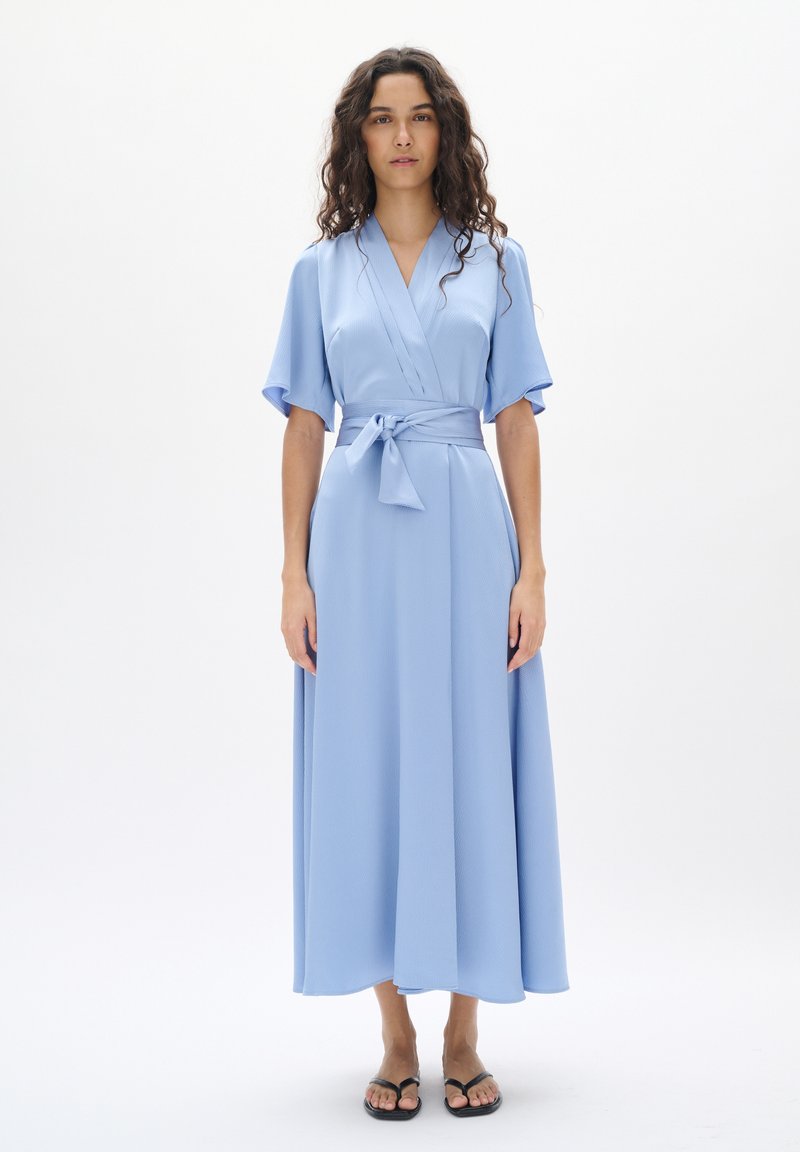 Helles blaues Wickelkleid mit kurzen, schwingenden Ärmeln, einem gebundenen Gürtel und einem bodenlangen Rock aus weichem, strukturiertem Stoff. Sandalen sind unten sichtbar.