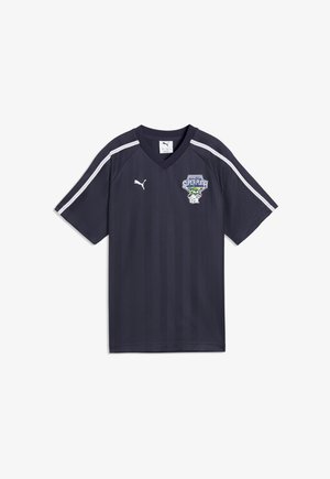 Ciemnogranatowa sportowa koszulka z dekoltem w serek, z białym logo Puma po prawej stronie klatki piersiowej, białe paski na ramionach oraz herb drużyny „Super Puma” po lewej stronie klatki piersiowej.