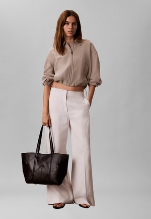 Frau trägt beige, kurz geschnittene Jacke mit Reißverschluss und weite hellrosa Hosen, hält große schwarze gesteppte Tragetasche mit Doppelgriffen.