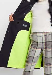 Abrigo negro con un forro interior verde neón, con un logo en el lado izquierdo. Lleva puesto sobre pantalones de cuadros grises con acentos rosas.