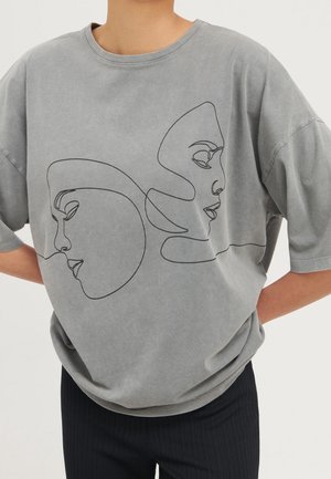 Szara oversize koszulka z czarną ciągłą liniową grafiką dwóch nałożonych na siebie kobiecych twarzy, noszona przez osobę z rękami za plecami.