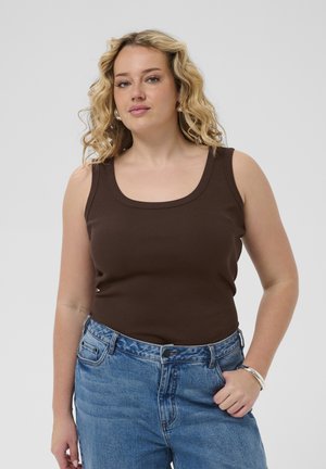 Kvinde med krøllet blondt hår iført en ærmeløs brun top og blå jeans, stående med den ene hånd i lommen mod en ensfarvet baggrund.