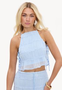Femme blonde portant un crop top plissé sans manches bleu clair et une jupe assortie avec de grandes boucles d'oreilles et un bracelet en or au poignet droit.