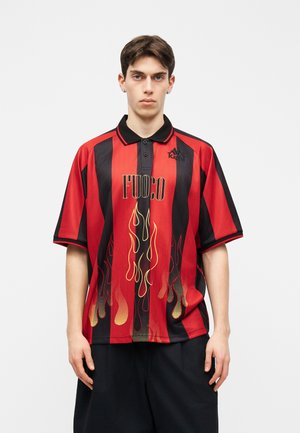 Νέος άνδρας που φοράει κόκκινο και μαύρο ριγέ πόλο με κίτρινα φλόγινα γραφικά και το κείμενο "FUOCO" στέκεται μπροστά σε λευκό φόντο.