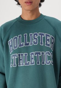 Felpa verde in morbido cotone, con la scritta "HOLLISTER ATHLETICS" in grandi caratteri bianchi e blu navy sul petto. Maniche raglan.