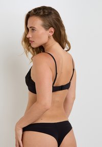 Giovane donna con capelli mossi, che indossa un reggiseno nero e una mutandina nera abbinata, posando di lato su uno sfondo chiaro e semplice.