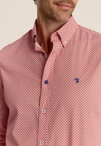 Korte mouwen blouse in het rood met een geometrisch patroon van witte diamanten. Voorzien van blauwe knopen en een klein logo op de borst.