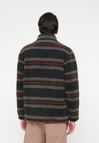 Ein Mann, der mit dem Rücken zur Kamera steht, trägt eine schwarz-grau gestreifte Fleecejacke und hellbraune Hosen vor einem schlichten weißen Hintergrund.