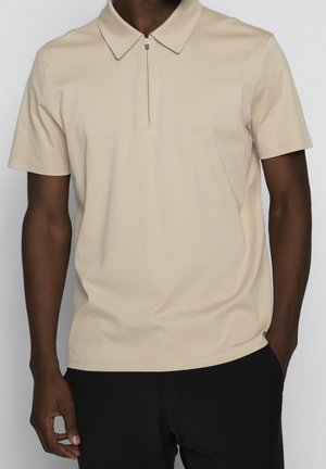 Short-sleeve polo shirt in beige, made from smooth fabric. Features a zippered collar and a fitted design, worn with black pants.