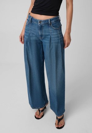 Kvinde iført løse blå denimjeans, sort ærmeløs top og sorte sandaler med åbne tæer, står mod en ensfarvet baggrund.