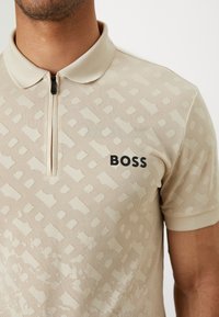 Béžové polo tričko s límečkem na zip, s texturovaným vzorem a černým logem "BOSS" na levém prsu. Krátké rukávy a prodyšná látka.