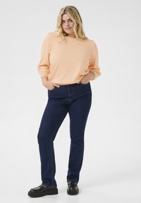 Jasno brzoskwiniowa, długosleeve bluzka z bufkami; w połączeniu z ciemnoniebieskimi, dopasowanymi dżinsami; modelka nosi czarne buty na platformie.