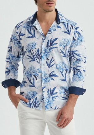 Chemise à boutons à motifs floraux avec des fleurs bleues et blanches sur un fond clair. Dotée d'un col et de poignets bleu marine. Fabriquée en tissu léger.