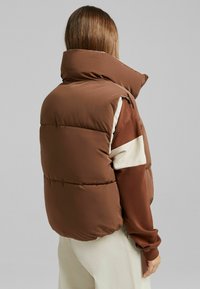 Gilet imbottito marrone con colletto alto, caratterizzato da dettagli crema e una texture lucida, progettato per il calore e il comfort.