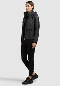 Femme portant une veste à capuche noire zippée avec des bandes blanches sur les manches, des leggings noirs et des baskets noires, debout de profil.