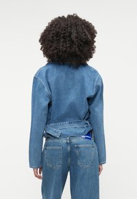 Laišuku jakas ar brīvu piegriezumu, garām piedurknēm un aizmugures saites detaļu, savienota ar atbilstošām augsto jostu džinsu biksēm. Zila krāsa visā.